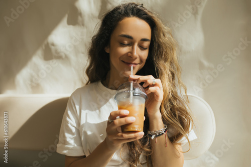 Young woman with long curly hair drinking iced coffee on beige background.