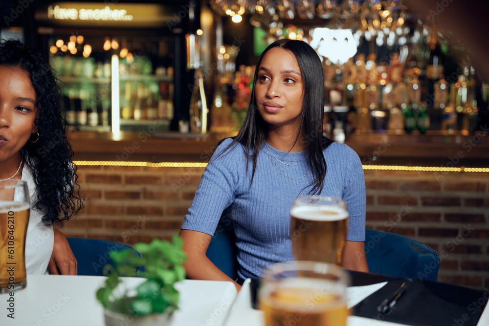 Young women in casual clothing looking away while sitting at table with beers and bill in restaurant