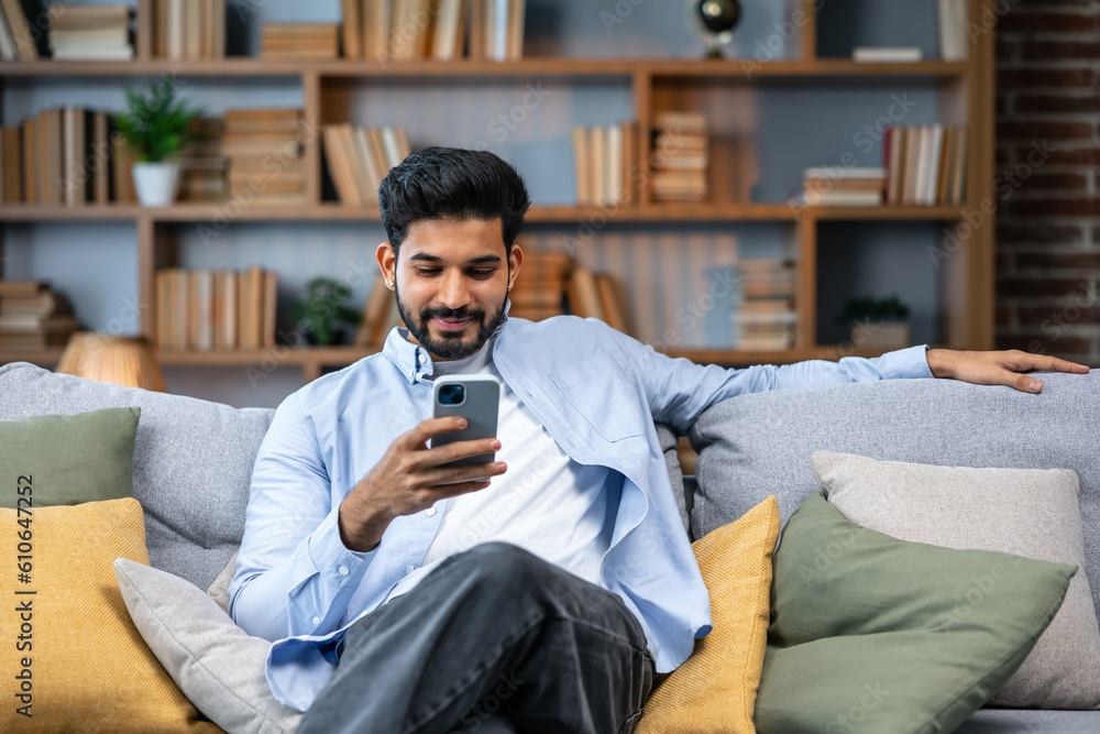 Young indian man holding smartphone tech device using cell phone apps at home. Bearded ethnic guy texting messages looking at smartphone checking social media, ordering online or browsing. Young indian man holding smartphone tech device using cell phone apps at home. Bearded ethnic guy texting messages looking at smartphone checking social media, ordering online or browsing.