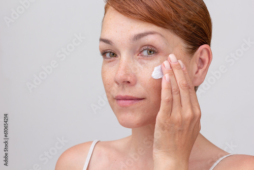 Portrait of cropped caucasian middle aged woman face with freckles and reddish hair applying cream on face on white background looking at camera