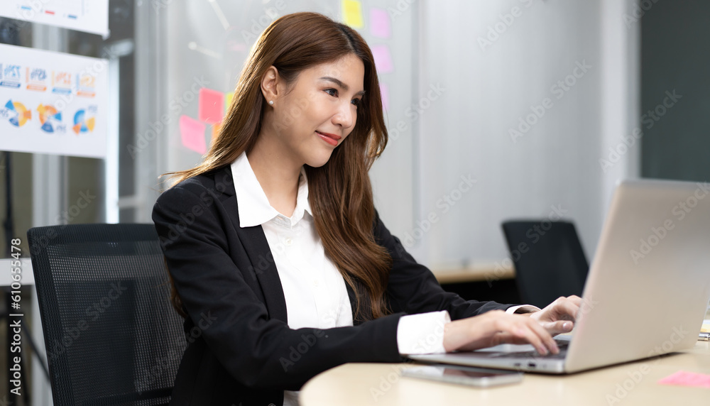 Young businesswoman sitting in office using notebook computer with smile. Smiling beautiful Asian professional executive manager happy working. Smart confident female entrepreneur at modern workplace Young businesswoman sitting in office using notebook computer with smile. Smiling beautiful Asian professional executive manager happy working. Smart confident female entrepreneur at modern workplace
