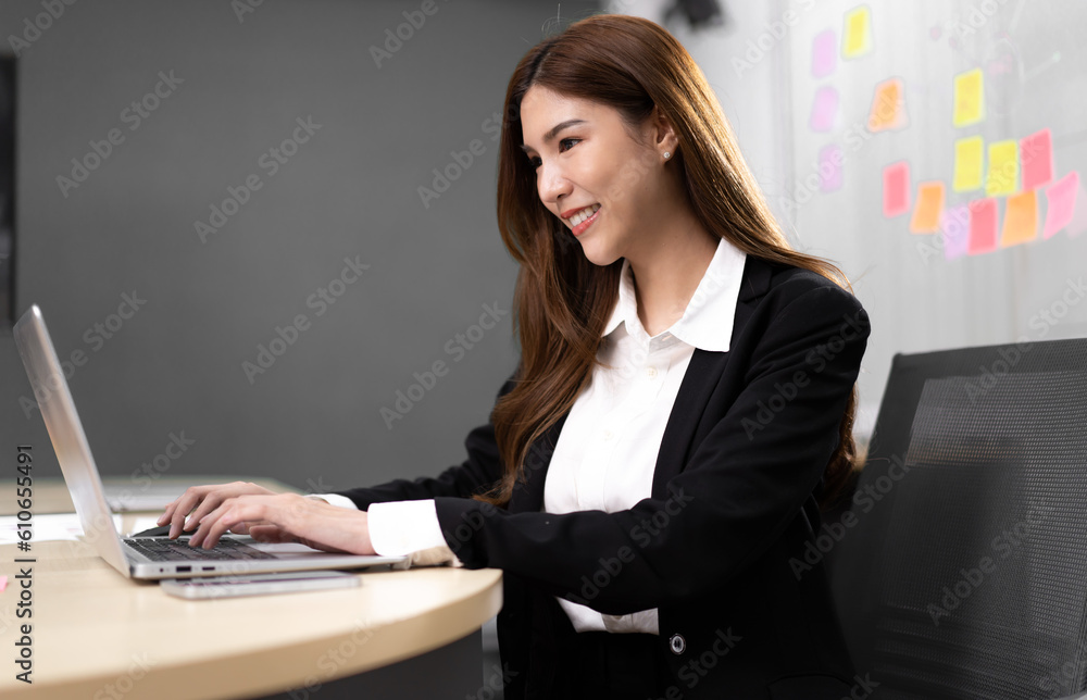 Young businesswoman sitting in office using notebook computer with smile. Smiling beautiful Asian professional executive manager happy working. Smart confident female entrepreneur at modern workplace Young businesswoman sitting in office using notebook computer with smile. Smiling beautiful Asian professional executive manager happy working. Smart confident female entrepreneur at modern workplace