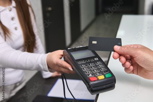 Close up manager administrator at reception counter holding out a reader POS terminal to unrecognizable client customer paying by black mockup credit card via NFC technology. People. Business. Finance