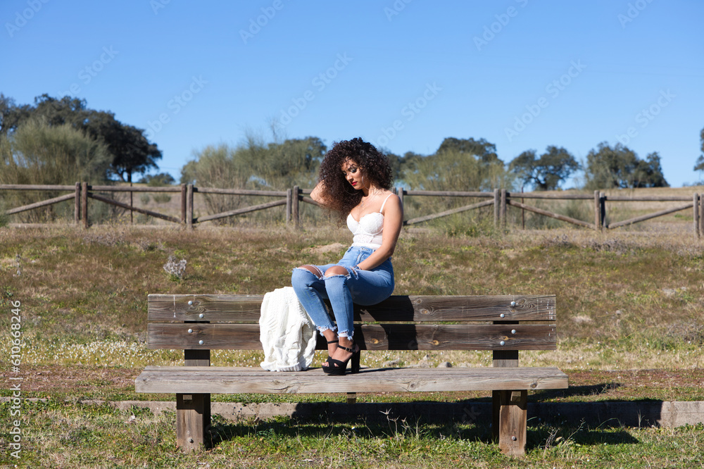 Spanish, brunette, curly-haired woman sits on a bench on the path leading to the forest. She is dressed in jeans and a white top and enjoys the sunny day.