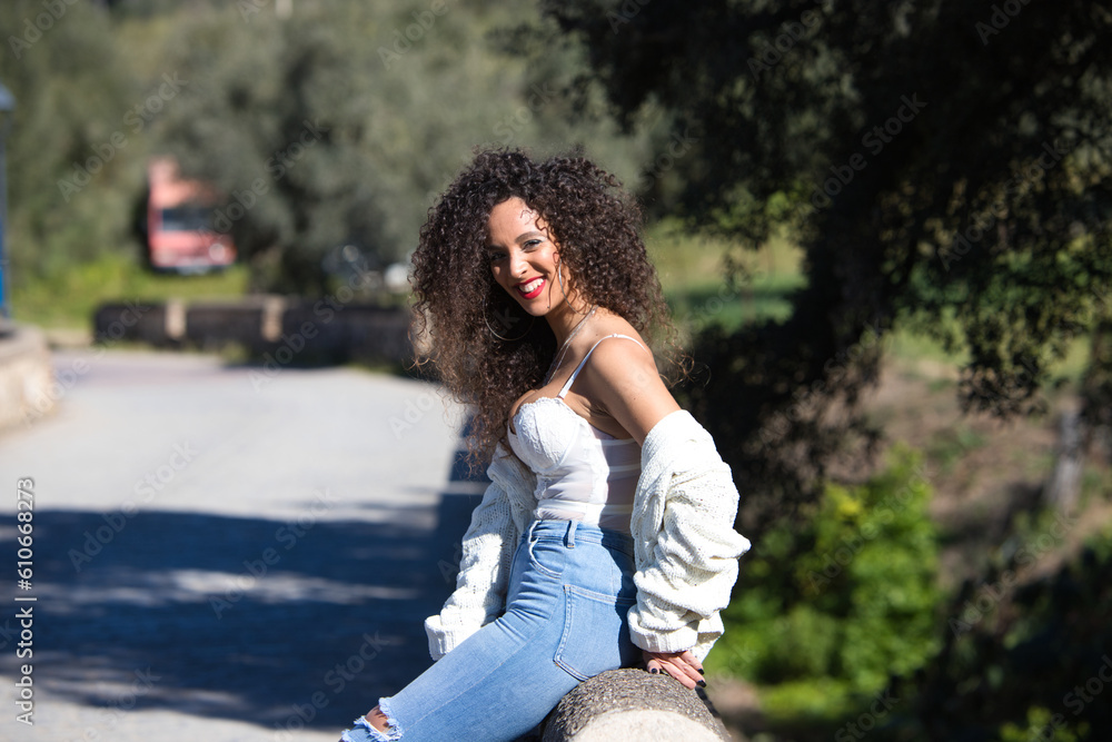 beautiful brunette woman with curly hair sitting on a stone wall in the forest. Trees and green forest vegetation in the background. The woman is happy and smiling. beautiful brunette woman with curly hair sitting on a stone wall in the forest. Trees and green forest vegetation in the background. The woman is happy and smiling.