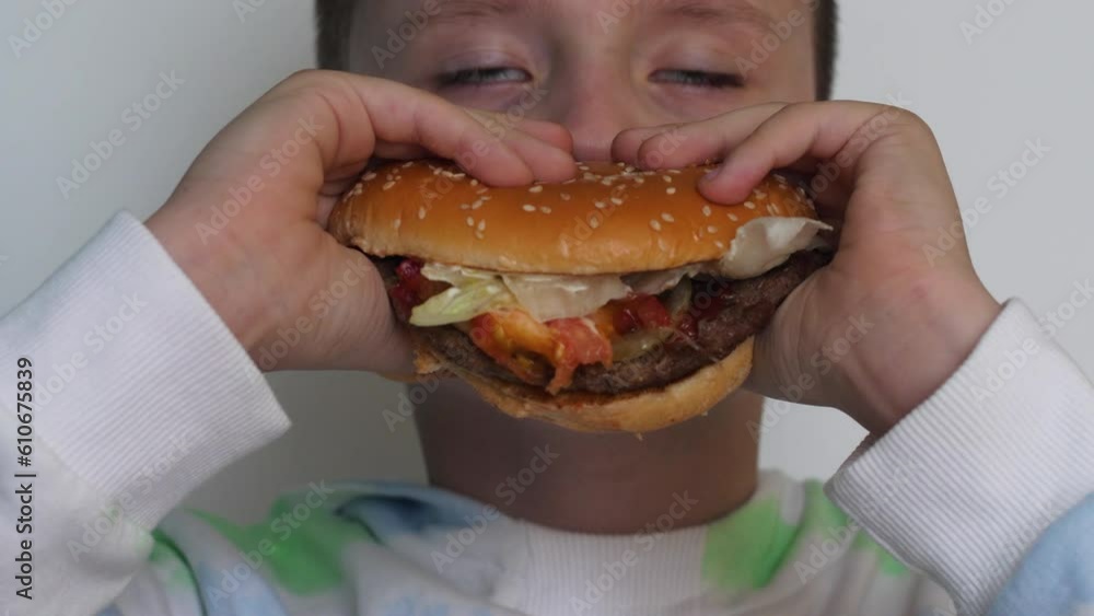 portrait of a caucasian boy hungry biting a huge burger, closing his eyes in enjoyment.european schoolboy has a snack in a school canteen, a cafe eats a hamburger portrait of a caucasian boy hungry biting a huge burger, closing his eyes in enjoyment.european schoolboy has a snack in a school canteen, a cafe eats a hamburger