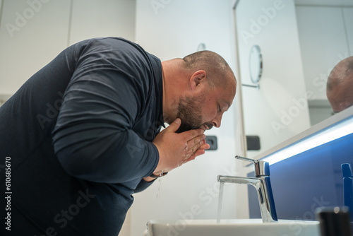 Fat, bald, bearded middle-aged man washes face in bathroom home int sink. Overweight and sweat. Daily routine, hygiene procedure, morning care and body cleanliness concept