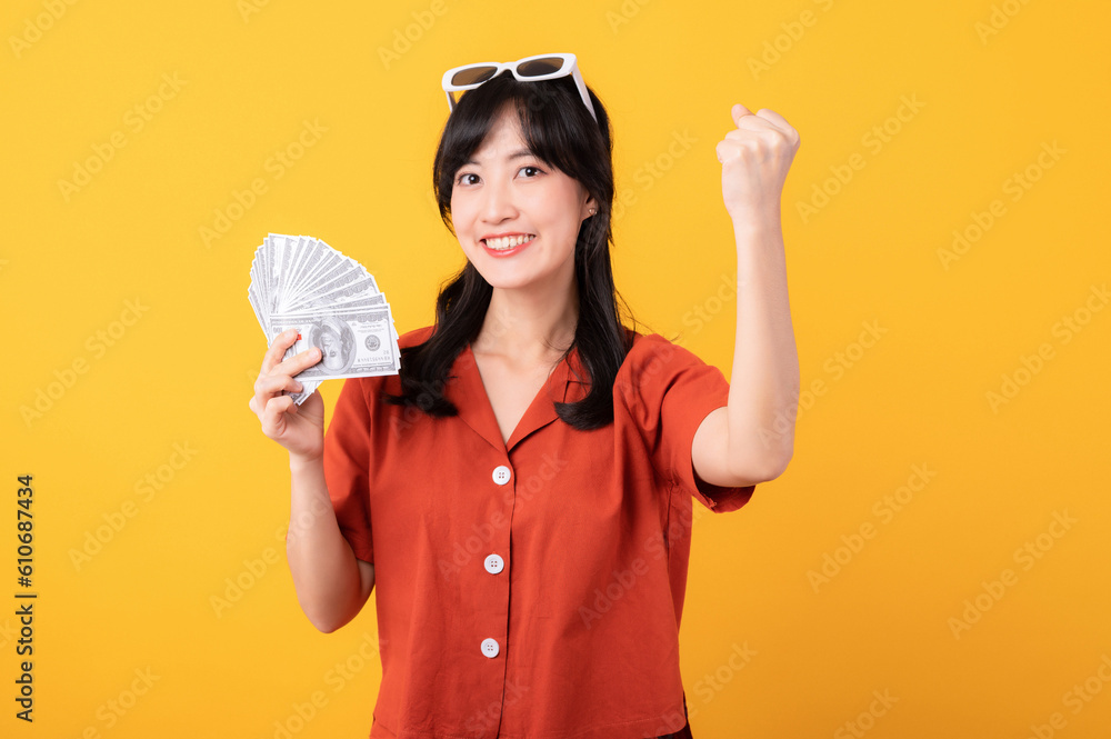 Portrait young beautiful asian woman happy smile dressed in orange clothes holding cash money and celebrate her success isolated on yellow background. Be rich concept. Portrait young beautiful asian woman happy smile dressed in orange clothes holding cash money and celebrate her success isolated on yellow background. Be rich concept.