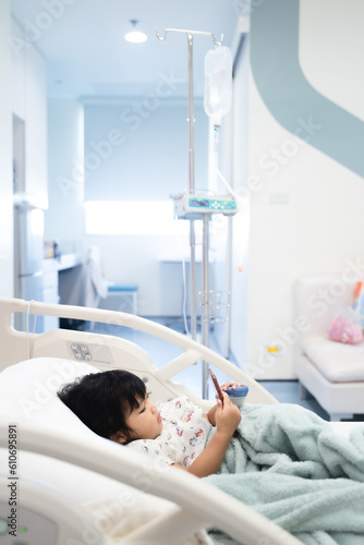 child is receiving medication through intravenous fluid therapy in hospital bed, focus on drip, and using smart phone
