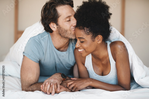 Happy interracial couple, bed and morning kiss in relax or bonding relationship at home. Man kissing woman on forehead in happiness or embrace for love or relaxing weekend together in the bedroom