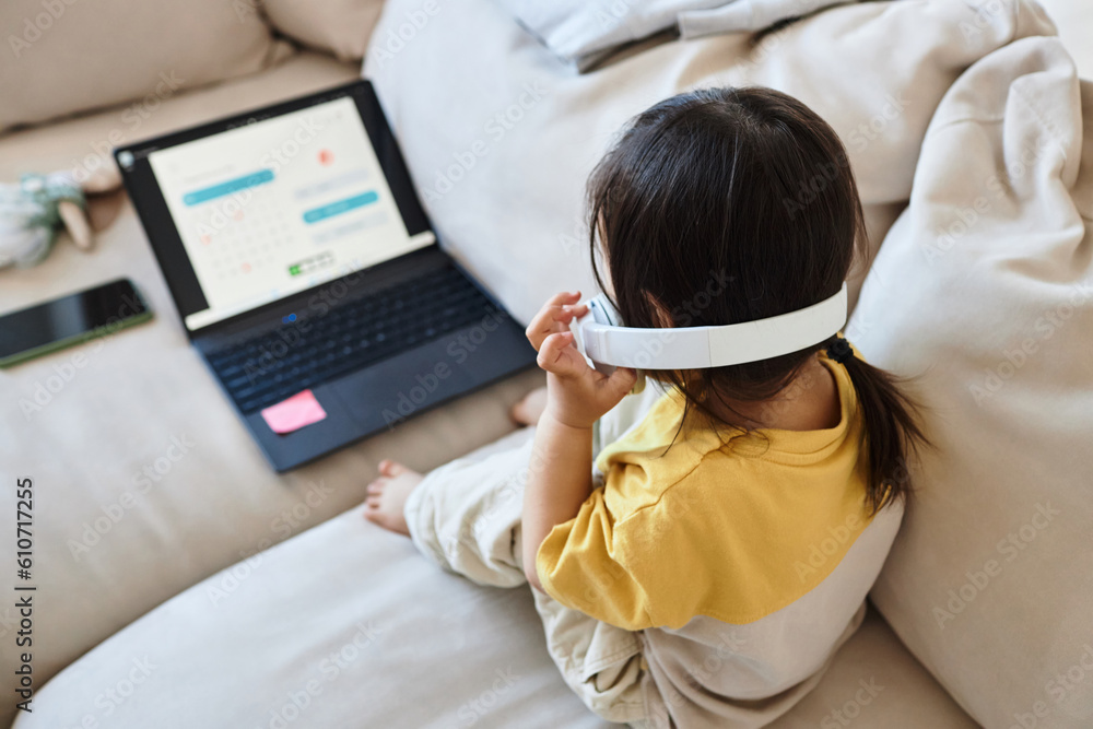 Rear view of little girl in wireless headphones sitting on sofa in ...