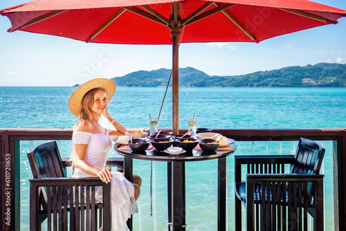 Young woman enjoys a delicious lunch in a luxury hotel resort. Girl sitting at a table with glass of water, looking out at the sea. Female in straw hat enjoy summer vacation. © TravelMedia