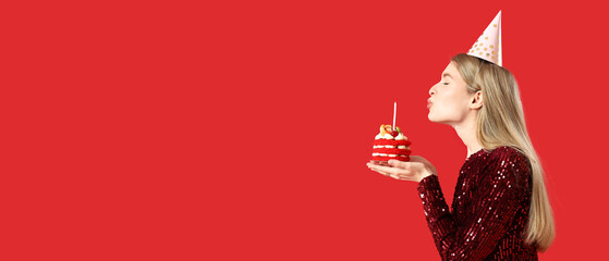 Beautiful young woman blowing out candle on tasty birthday cake against red background with space for text
