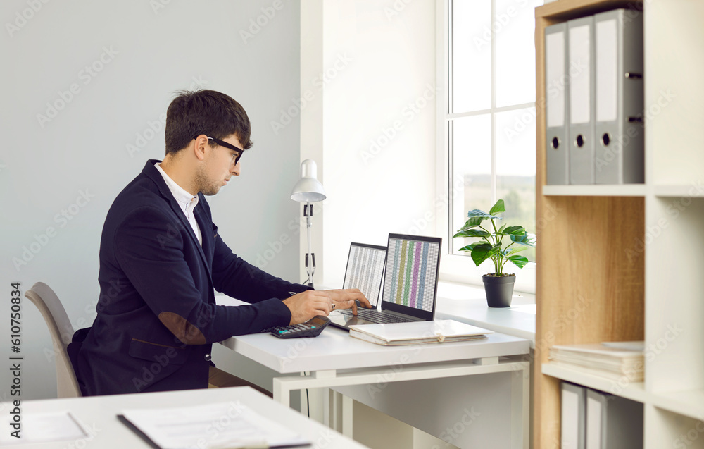 Professional financial accountant working in the office. Man in a suit and glasses sitting at his desk in front of laptop computers and looking at figures in digital business spreadsheets on screens Professional financial accountant working in the office. Man in a suit and glasses sitting at his desk in front of laptop computers and looking at figures in digital business spreadsheets on screens