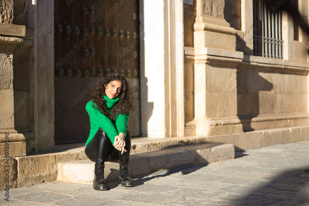 Young, beautiful, brunette woman with curly hair and a green turtleneck sweater, leather pants and black boots, sitting on a step, calm and relaxed. Concept beauty, fashion, autumn, winter, cold.