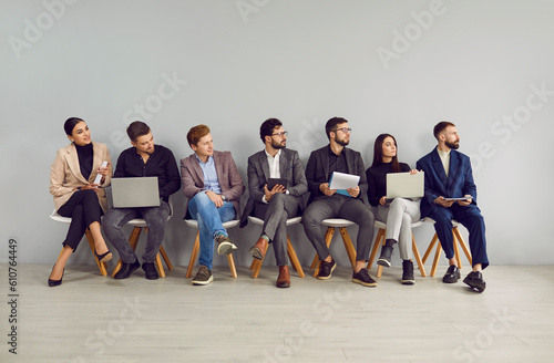 People waiting for a job interview. Group of young men and women with CVs and laptops sitting in line by a gray office wall and looking aside at the door that is out of our sight