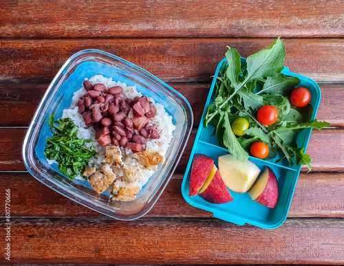 
lucnch box with tiica Brazilian food with rice, beans and chicken and zucchini and another one with rocket and tomatoes and apple salad on rustic wooden background