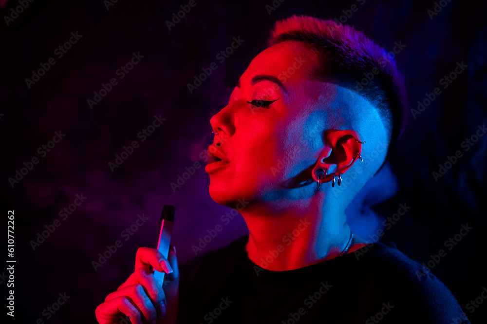 Fototapeta premium Portrait asian woman with shaved temples smokes vape in neon light.