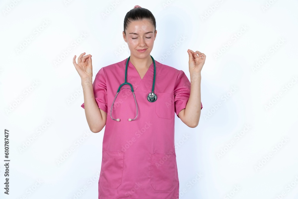 young caucasian doctor woman wearing pink uniform over white background doing yoga, keeping eyes closed, holding fingers in mudra gesture. Meditation, religion and spiritual practices. young caucasian doctor woman wearing pink uniform over white background doing yoga, keeping eyes closed, holding fingers in mudra gesture. Meditation, religion and spiritual practices.
