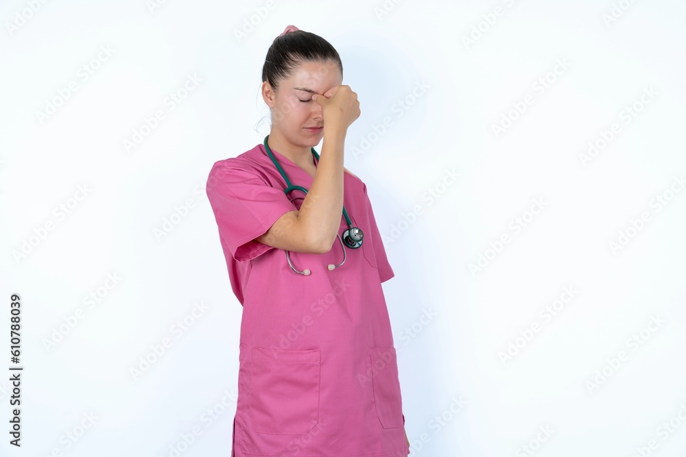 Very upset, young caucasian doctor woman wearing pink uniform over white background touching nose between closed eyes, wants to cry, having stressful relationship or having troubles with work Very upset, young caucasian doctor woman wearing pink uniform over white background touching nose between closed eyes, wants to cry, having stressful relationship or having troubles with work