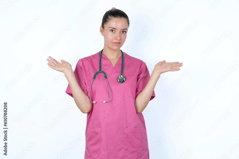 Puzzled and clueless young caucasian doctor woman wearing pink uniform over white background with arms out, shrugging shoulders, saying: who cares, so what, I don't know. Negative human emotions. Puzzled and clueless young caucasian doctor woman wearing pink uniform over white background with arms out, shrugging shoulders, saying: who cares, so what, I don't know. Negative human emotions.