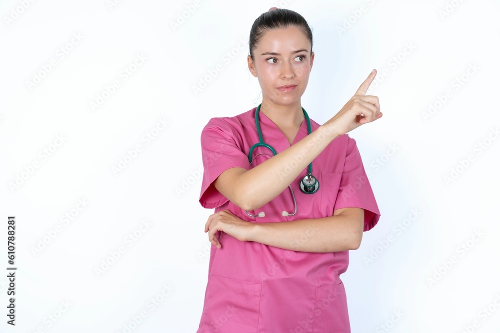 Portrait of young caucasian doctor woman wearing pink uniform over white background posing on camera with tricky look, presenting product with index finger. Advertisement concept. Portrait of young caucasian doctor woman wearing pink uniform over white background posing on camera with tricky look, presenting product with index finger. Advertisement concept.