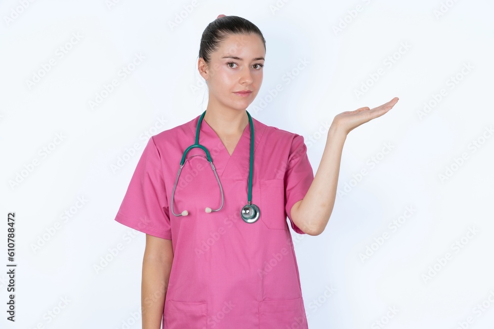 Positive glad young caucasian doctor woman wearing pink uniform over white says: wow how exciting it is, has amazed expression, shows something on blank space with open hand. Advertisement concept. Positive glad young caucasian doctor woman wearing pink uniform over white says: wow how exciting it is, has amazed expression, shows something on blank space with open hand. Advertisement concept.