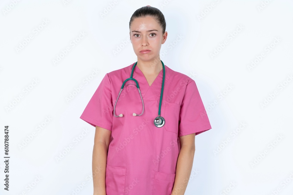 Gloomy, bored young caucasian doctor woman wearing pink uniform over white background frowns face looking up, being upset with so much talking hands down, feels tired and wants to leave. Gloomy, bored young caucasian doctor woman wearing pink uniform over white background frowns face looking up, being upset with so much talking hands down, feels tired and wants to leave.