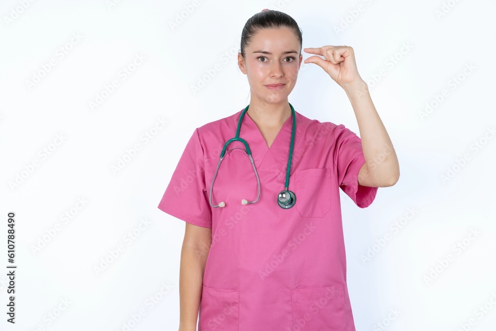 Upset young caucasian doctor woman wearing pink uniform over white background shapes little gesture with hand demonstrates something very tiny small size. Not very much Upset young caucasian doctor woman wearing pink uniform over white background shapes little gesture with hand demonstrates something very tiny small size. Not very much