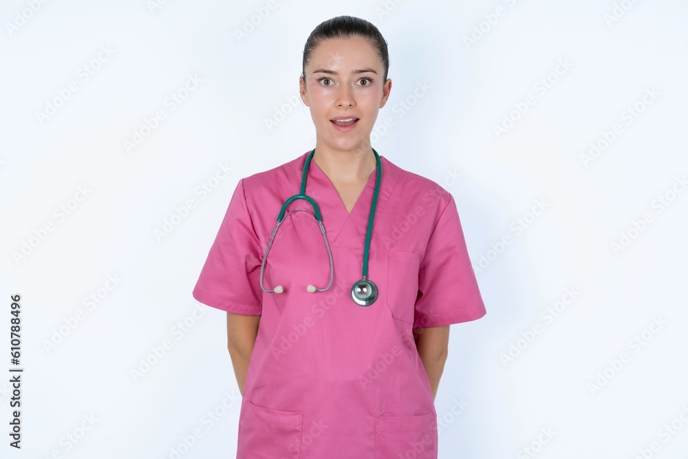 Emotional attractive young caucasian doctor woman wearing pink uniform over white with opened mouth expresses great surprise and fright, stares at camera. Unexpected shocking news and human reaction. Emotional attractive young caucasian doctor woman wearing pink uniform over white with opened mouth expresses great surprise and fright, stares at camera. Unexpected shocking news and human reaction.