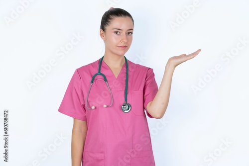 Positive glad young caucasian doctor woman wearing pink uniform over white says: wow how exciting it is, has amazed expression, shows something on blank space with open hand. Advertisement concept.