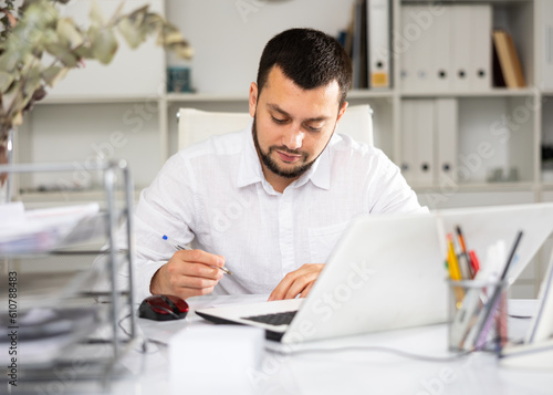 European man office worker sitting at table in front of his laptop and making notes on paper.