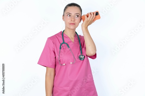 Slika na platnu Smiling young caucasian doctor woman wearing pink uniform over white background listening a voice message from her smartphone