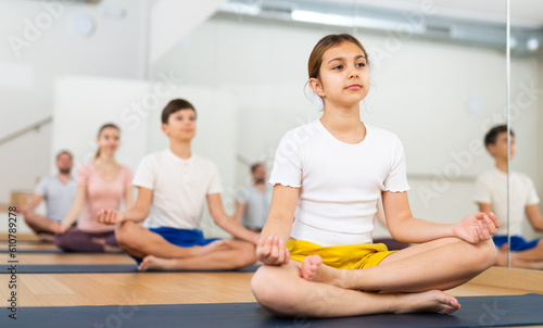 Portrait of focused teen girl meditating in yoga position Padmasana during family training at gym..