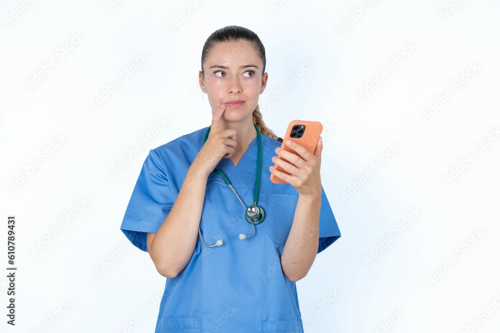 young caucasian doctor woman wearing medical uniform over white background thinks deeply about something, uses modern mobile phone, tries to made up good message, keeps index finger near lips. young caucasian doctor woman wearing medical uniform over white background thinks deeply about something, uses modern mobile phone, tries to made up good message, keeps index finger near lips.