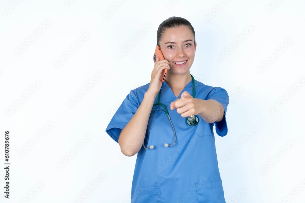 Positive young caucasian doctor woman wearing medical uniform over white background indicates directly at camera has telephone conversation smiles broadly enjoys talking long hours. You join me Positive young caucasian doctor woman wearing medical uniform over white background indicates directly at camera has telephone conversation smiles broadly enjoys talking long hours. You join me