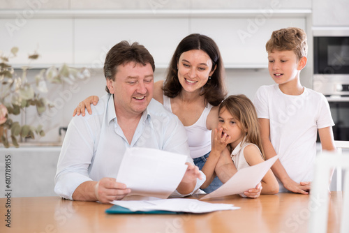 Positive husband is sitting at kitchen and reading document about career advancement at work, moneymaking financial report. Smiling wife and happy children hug successful dad