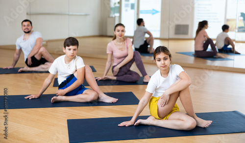 Positive preteen girl doing yoga with brother and parents in fitness studio, sitting in twisting asana Matsyendrasana ..