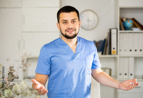 Portrait of positive man physician wearing surgical scrubs standing in his office and explaining with gestures.