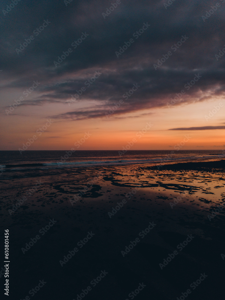 Sunset at the beach. The sky is orange and blue, with pink and purple clouds. Waves crash on the shoreline, reflecting the sun on the wet sand.