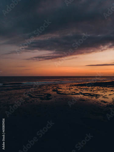 Sunset at the beach. The sky is orange and blue, with pink and purple clouds. Waves crash on the shoreline, reflecting the sun on the wet sand.
