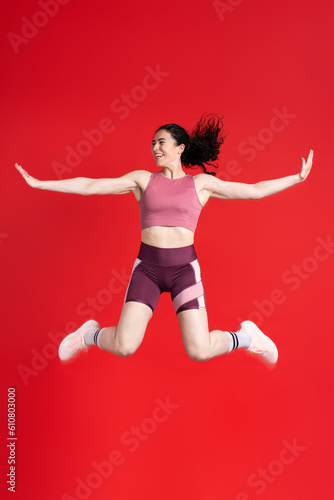 Sporty athletic woman doing gym exercises, jumping high, isolated on red background