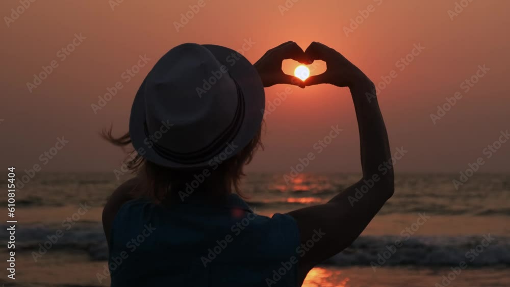 Back view of a young woman wearing hat making heart shaped hands against the beautiful sunset on the sea. Female silhouette on the setting sun background.