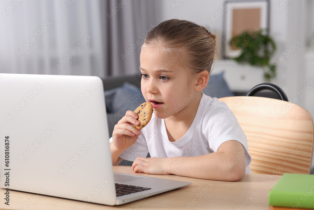 Little girl using laptop and eating biscuit at table indoors. Internet addiction Little girl using laptop and eating biscuit at table indoors. Internet addiction
