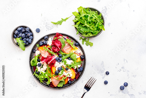 Gourmet fresh salad with arugula, radicchio, sweet peaches, ham, cheese and blueberries. White table background, top view