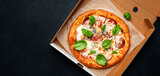 Hot pizza in box with spicy salami sausage, mozzarella cheese, tomato sauce and green basil, just delivered, black table background, top view, copy space banner
