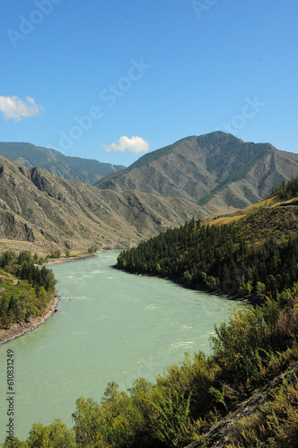 Bends of a broad turquoise river flowing through a valley at the foot of a high mountain range on a sunny summer day.