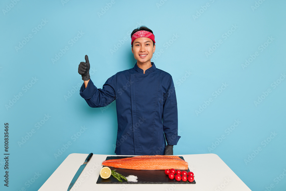 Friendly young oriental cook in blue uniform stands near table with cutting board with salmon fillet, knife and products for making sushi, tasty food concept, copy space Friendly young oriental cook in blue uniform stands near table with cutting board with salmon fillet, knife and products for making sushi, tasty food concept, copy space