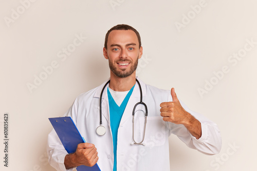 Friendly young male doctor with beard stands on white background wearing white coat with stethoscope on his neck and clipboard in hand  thumb up and smiles  professional people concept  copy space