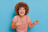 Happy cute girl with afro hairstyle wearing polka-dot top, stands pressing hand to heart laughs with eyes closed, fun time concept, copy space